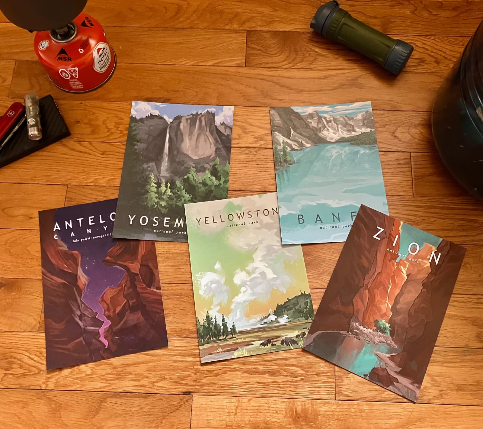 Photo mockup of printed National Park poster postcards for Zion, Yosemite, Yellowstone, Banff, and Antelope Canyon laid out on a wood floor next to camping gear