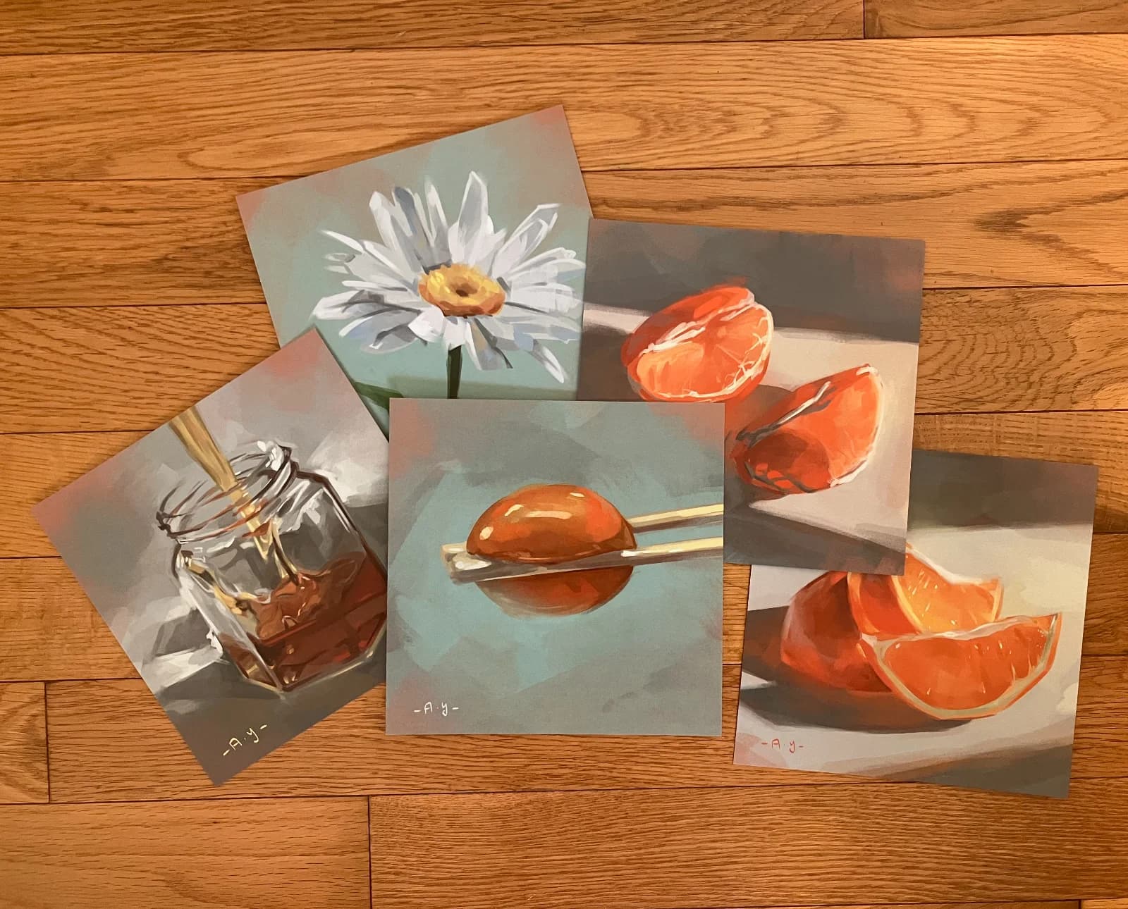 Photo mockup of printed still life art prints — honey, a daisy, a kumquat, and citrus slices — laid out on a wood floor